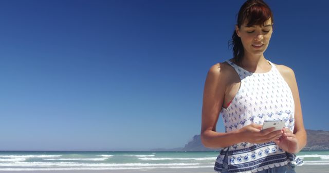 Young woman standing on sandy beach, looking at smartphone. Blue ocean waves and clear sunny sky. Ideal for content on summer vacations, technology use outdoors, leisure, or coastal trips.