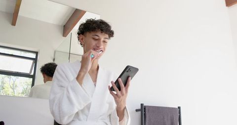 Young Man Brushing Teeth While Using Smartphone in Modern Bathroom