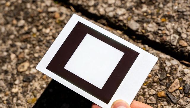Hand holding calibration target black square on white card casting shadow on sunlit pavement