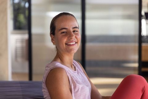 Smiling woman sitting on low bed in pink activewear, relaxing at home with frosted glass