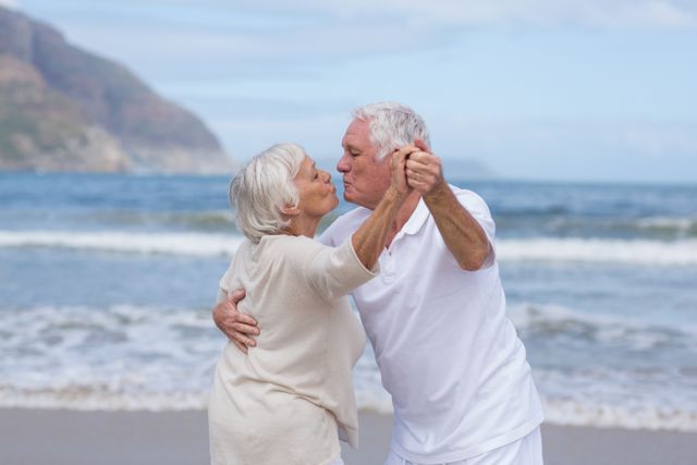 Senior couple enjoying a romantic moment at the beach, dancing and kissing by the ocean. Ideal for use in advertisements promoting retirement plans, senior living, vacation destinations, or lifestyle blogs focusing on love and relationships in later life.
