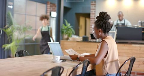 African woman working remotely at communal cafe table with laptop and documents