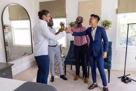 Diverse male friends toasting with champagne in stylish bedroom suite before wedding celebration