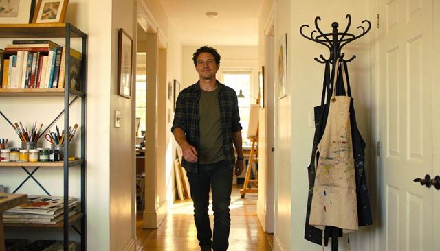 Male artist walking through bright home studio hallway with paint-splattered apron
