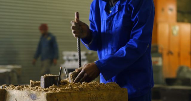 Worker in blue uniform molding industrial parts with sand in a factory workshop. The environment is characterized by industrial machinery and rugged workspace. Excellent visual for topics related to heavy industry, manufacturing processes, craftsmanship, industrial training, and labor-intensive jobs.