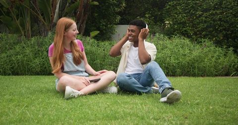 Diverse young couple sitting on lawn, woman holding smartphone, man wearing headphones, smiling