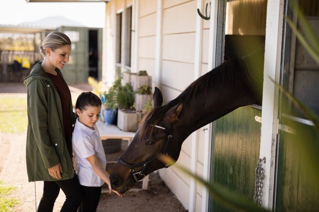 Mother and daughter enjoying time together feeding a horse in a stable. Ideal for promoting family bonding activities, equestrian centers, rural lifestyle, and animal care. Can be used in advertisements, blogs, and articles about parenting, outdoor activities, and farm life.