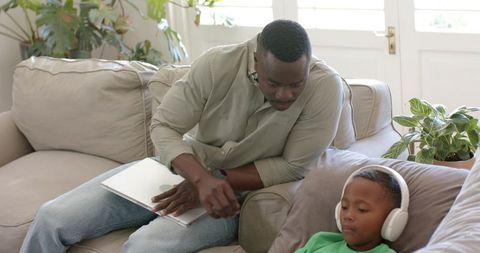 African american father bonding with son on couch with laptop, child listening with headphones