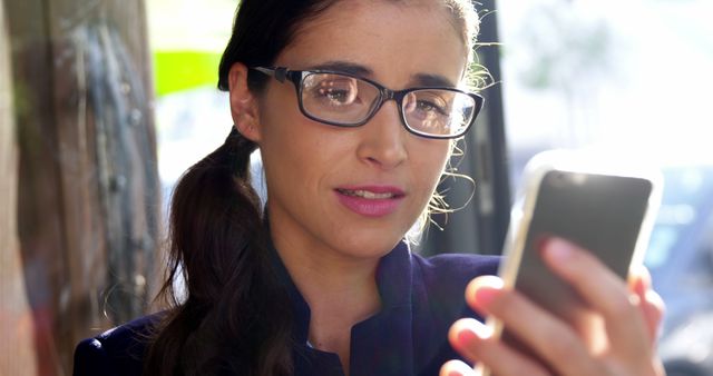 A woman wearing glasses uses her smartphone outdoors in an urban environment. She appears focused on the screen, suggesting engagement with technology and connectivity. This can be used in contexts relating to communication, mobile technology, city life, and women in tech.