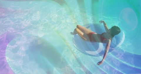 Sunlit woman reclining on tube floating over turquoise pool steps with pastel lens flare