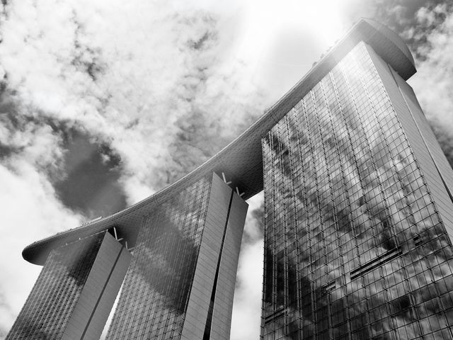 The image captures a striking high-rise building with a reflective glass facade under a cloudy sky, presented in black and white. The futuristic design of the structure stands out, showcasing contemporary architecture. This could be effectively used in projects related to urban development, architecture, real estate, and modern cityscapes.