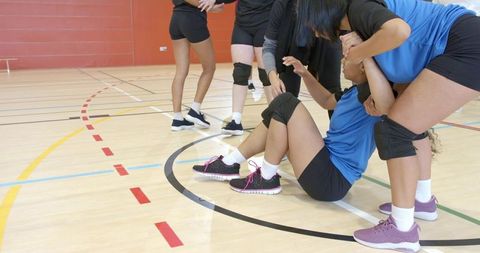Helping teammate sitting on gym court by diverse female athletes showing team support