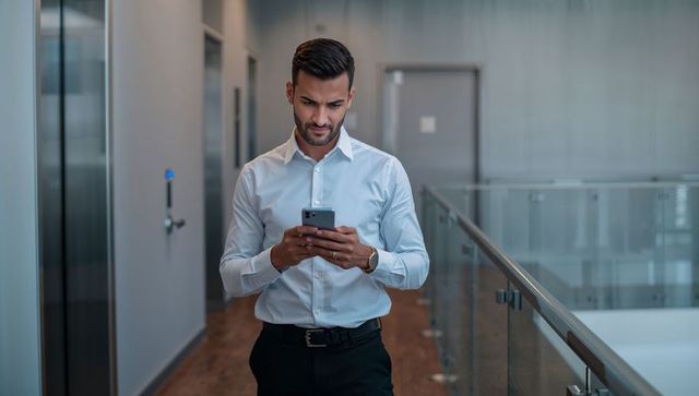 Businessman walking office corridor checking smartphone by glass railing