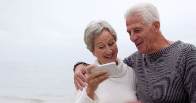 Senior couple embracing and smiling while looking at a smartphone on a beach. Suitable for themes related to love, retirement, technology use among seniors, leisure activities, and family bonding.