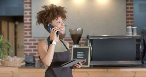 Smiling barista talking on smartphone while holding tablet behind espresso machine in coffee shop
