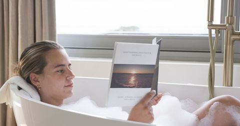 Woman relaxing in freestanding tub reading sunset-cover paperback amid bubble bath