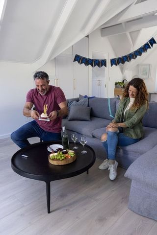 Couple celebrating birthday on loft sofa, man holding cake with candles, cozy home celebration