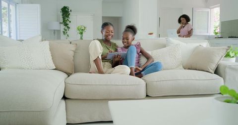 African american sisters sharing tablet on beige sectional sofa in sunlit modern home