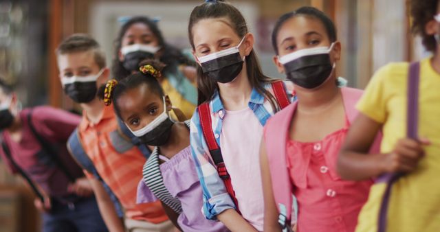 Group of children standing in a line indoors, wearing face masks and backpacks, representing school routines during the pandemic. The image is suitable for educational, health, and safety campaigns, back-to-school promotions, and diversity advocacy in educational settings.