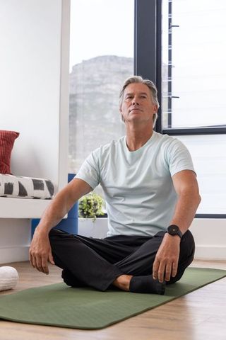 Mature man practicing indoor meditation and gentle stretch on yoga mat by window