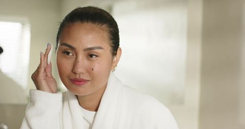 Mid-adult asian woman applying skincare with cotton pad wearing white bathrobe