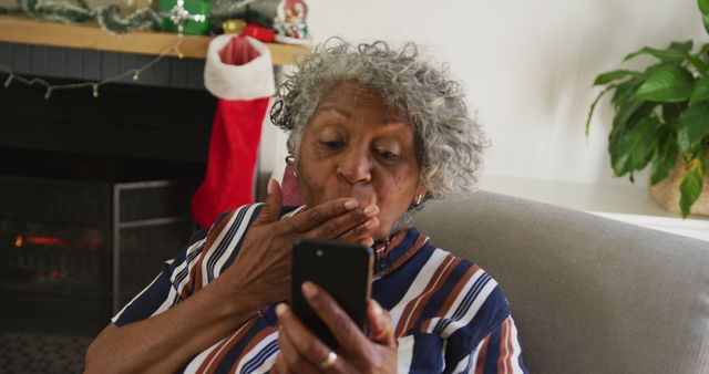Elderly woman sitting on a couch, looks surprised while holding smartphone in hand. Scene captures cozy Christmas ambiance with lit fireplace, decorative stocking, and festive decorations. Ideal for use in articles about seniors embracing technology, holiday marketing for elderly products, or internet safety tips for senior citizens.