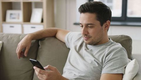 Young man relaxing on sofa using smartphone for messaging, browsing and remote work at home
