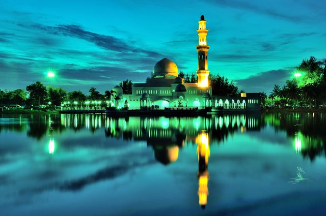 This image features a stunning mosque alongside a calm lake during dusk. The beautifully illuminated mosque and its reflection in the tranquil water create a symmetrical and serene atmosphere. Ideal for use in travel brochures, religious publications, websites about architecture, backgrounds for inspirational quotes, and emphasizing tranquility in advertising materials.