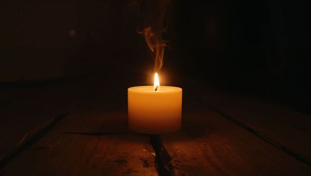 Warm glowing pillar candle burning on worn wooden planks with rising smoke and melted wax
