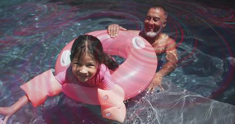 Smiling Girl Floating on Pink Donut, Father Supporting Her While Playing in Sunny Pool
