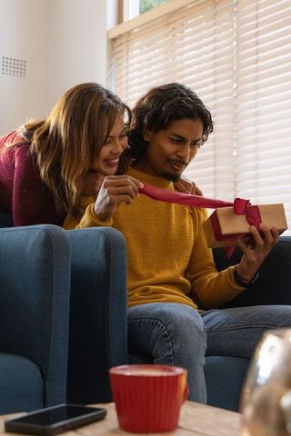 Man receiving surprise gift on couch while partner playfully pulling ribbon