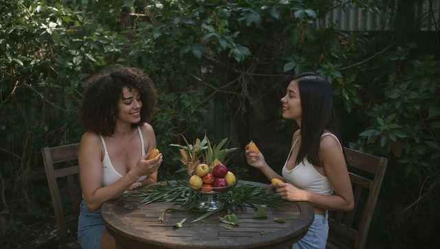 Two women chatting in backyard sharing tropical fruit on wooden patio table wearing tank tops