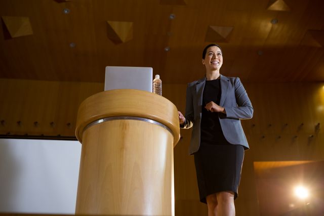 Confident female executive giving a speech at a conference center. Ideal for use in articles about leadership, business presentations, corporate events, public speaking, and professional development.