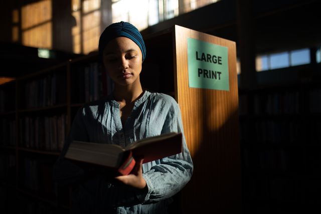 Front view of a biracial female student wearing a dark blue hijab studying in a library, standing and holding a book in hands, reading it.