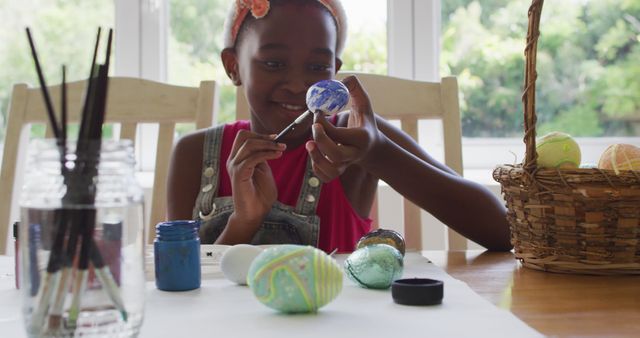 Young girl painting Easter eggs at a table. She is focused on adding details to one egg while a collection of painted eggs and painting supplies are in front of her. Ideal for use in Easter celebrations advertisements, children’s activity promotions, or creative arts and crafts content.
