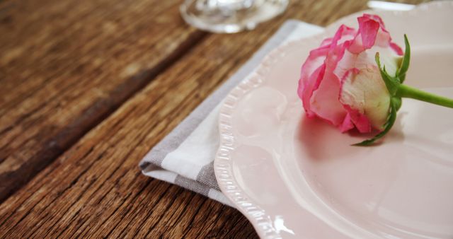A pink rose rests on an elegant plate beside a folded napkin, with copy space. The setting suggests a romantic or celebratory occasion, a fine dining experience.