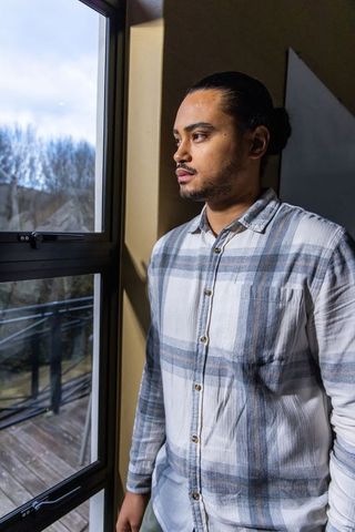 Contemplative man standing by tall window wearing light plaid shirt in sunlit interior