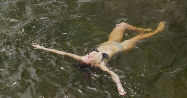 Young woman is seen floating peacefully on her back in a river, enjoying a relaxing summer day outdoors. Appropriate for use in promoting summer activities, travel, leisure, tranquil outdoor retreats, personal freedom, and lifestyle imagery. Ideal for websites, blogs, and marketing materials focused on nature, wellness, and relaxation.
