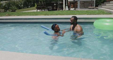 African american father teaching teenage son to swim in backyard pool on sunny day with tube