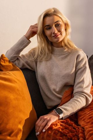 Woman relaxing on sofa wearing beige knit sweater and rust-orange throw, velvet cushion