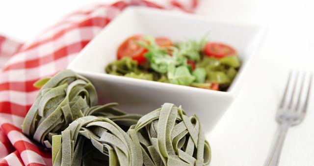 Delicious fresh spinach tagliatelle served with cherry tomatoes and arugula salad, placed beside a checkered cloth and a fork. Perfect for illustrating healthy eating habits, vegetarian recipes, and Italian cuisine. Ideal for food blogs, recipe websites, and cooking magazines.