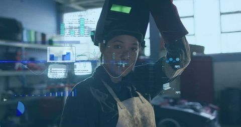 Woman welder lifting helmet with AR overlays in garage workshop wearing leather apron