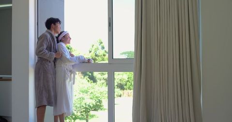 Couple relaxing and gazing through window in robes overlooking green garden