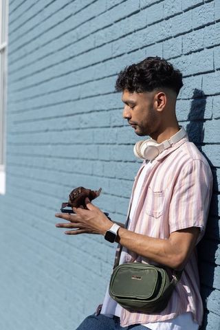 Indian man leaning on blue brick wall holding smartphone and pastry wearing headphones