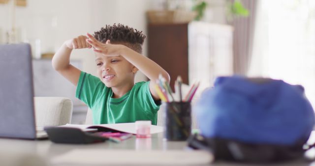A mixed-race boy sits at a desk with a laptop, attending an online class. He is raising his hands, indicating a feeling of engagement and enjoyment in learning. Color pencils, notebooks, and other study materials are scattered on his desk, demonstrating a vibrant educational setting at home. This image is perfect for use in educational content, online learning platforms, remote schooling, and digital educational tools promotion.
