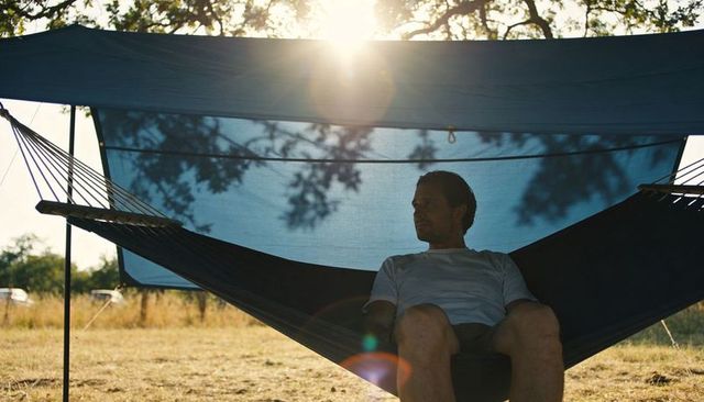Man resting in hammock under blue tarp sunshade at campsite meadow during golden hour