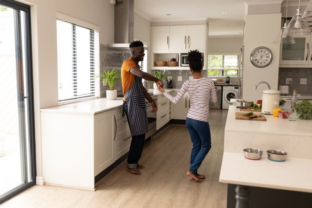 This image depicts a joyful African American couple dancing in a modern kitchen at home. The man is wearing an apron, suggesting they are cooking together. This image can be used for promoting lifestyle blogs, relationship advice articles, home decor websites, or advertisements focusing on domestic happiness and togetherness.