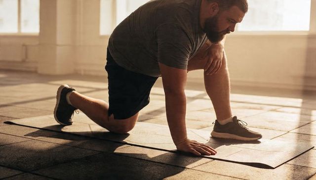 Bearded man kneeling and stretching knee on mat in sunlit gym studio for mobility