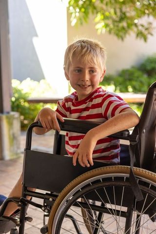 Blond boy sitting in manual wheelchair smiling on sunlit porch wearing red striped shirt