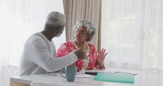 Senior man and woman sitting at dining table, engaging in serious discussion with papers and documents in front of them. Ideal for depicting communication among elderly, handling personal matters, or morning routines. Could be used in articles about relationships, financial planning, or health consultations for seniors.